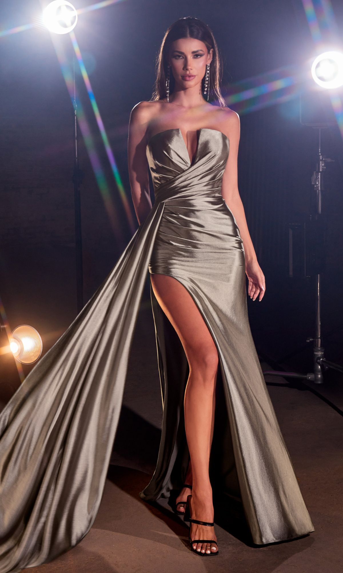 A woman poses confidently under studio lights in the Side-Drape Strapless Long Formal Dress CDS441, a silver floor-length gown with a high slit and dramatic drape, styled with black high-heeled sandals.